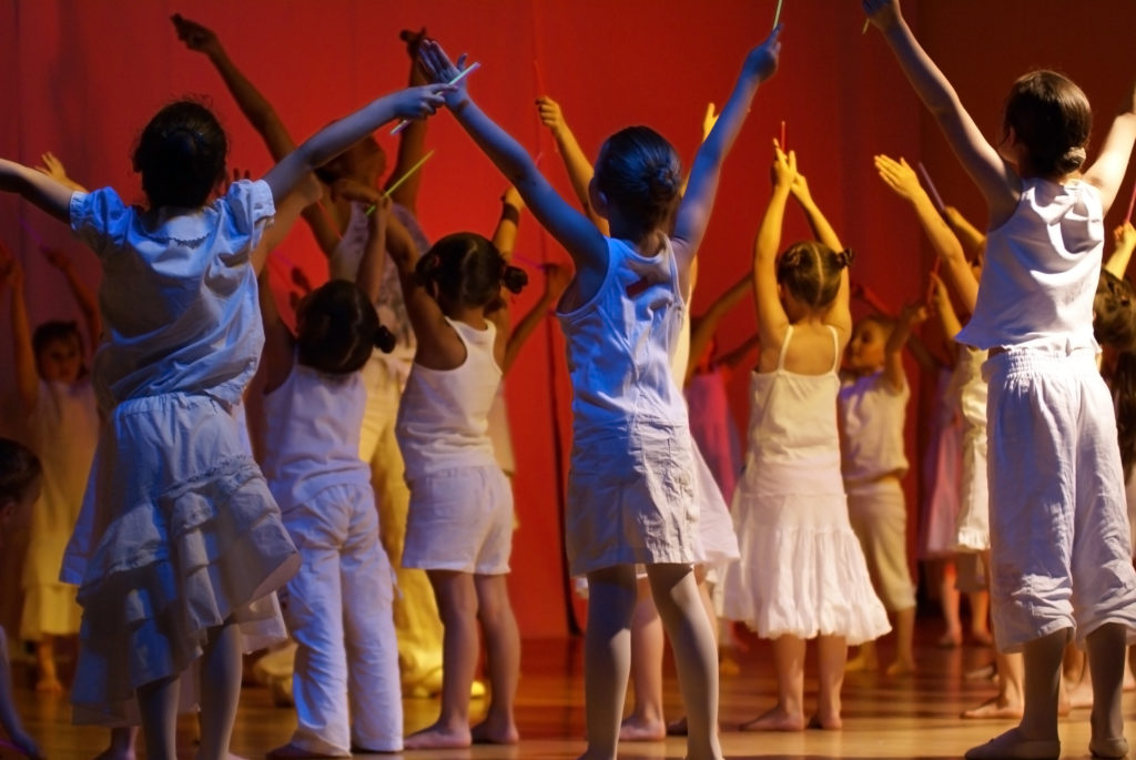 This photo featured a group of children on an indoor stage performing a dance. The children wear white clothing, some in skirts, shorts and short pants and with sleeveless and short-sleeved shirts. The photo captures them with raised arms and in a circle around their instructor. The children posed in the foreground have their backs to the audience and the photographer. They are holding small, colored wands. Credit: Adobe Stock Photo.