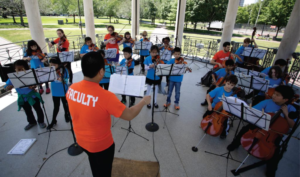 This photograph features a youth string orchestra performing outdoors in a covered park amphitheater. The background of the photograph shows the park's greenspace with manicured lawns and trimmed trees. In the amphitheater, youth are positioned in rows based on musical instrument and are angled around the conductor in a semi-circle. The conductor is in the foreground of the photo with back to the camera. Youth are wearing blue t-shirts, and the conductor and performing staff are wearing orange t-shirts. Photo Credit: Courtesy of EdVestors.