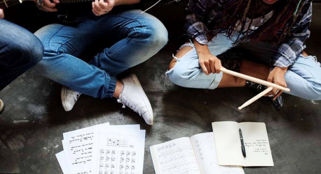 Image of people seated on the ground with their legs crossed. The image only shows the legs of the individuals, all three of whom are wearing blue jeans. Two figures are holding guitars, while the third is holding drum sticks. . There are notebooks and music paper in front of them.