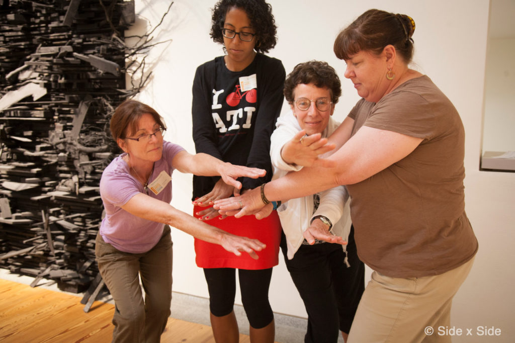 Four adults stand side-by-side, leaning in to overlap their hands in a SxS activity.