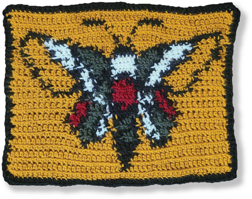 Crochet fiber arts piece of a moth. The moth is green, red, white and black and is set against a yellow background.