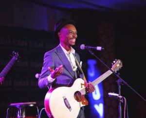 Photograph of Lawi (Francis Phiri). Francis is a Black man wearing a suit. He is holding a guitar and smiling while standing at a microphone.