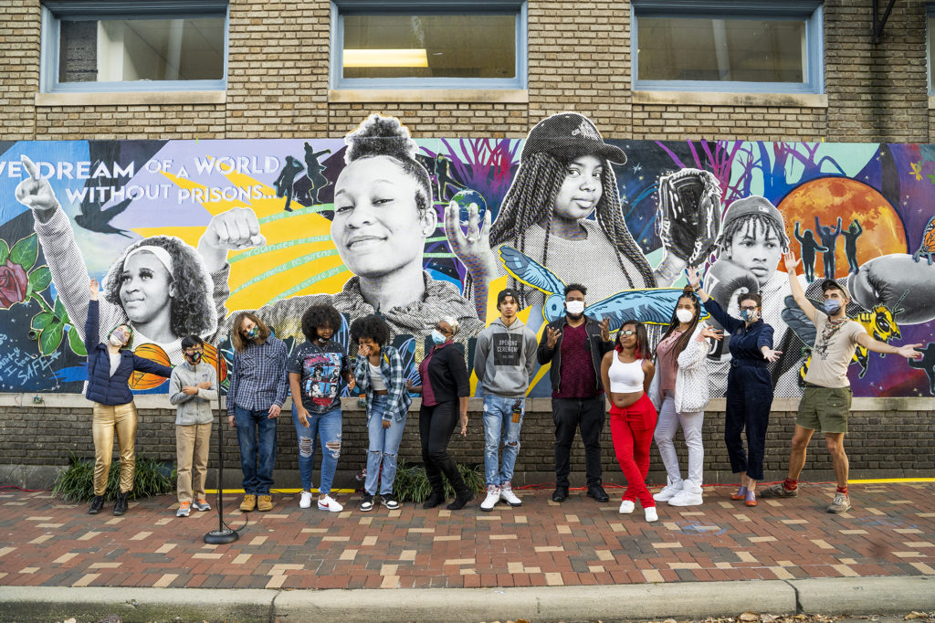Twelve youths stand in front of a colorful mural wall, laughing and showing off the art. The mural has youth faces and bright colors, with the words "we dream of a world without prisons..."