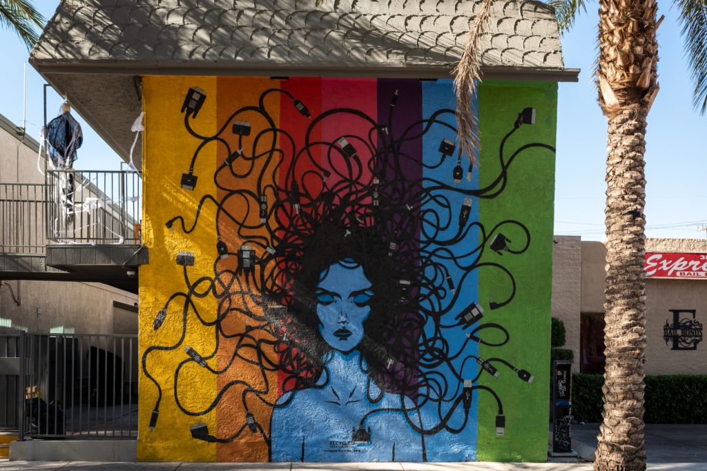 The photo captures an outdoor mural in the Las Vegas, Nevada, Arts District during a bright, sunny day. The artist painted the mural on the side of what appears to be an apartment complex. The mural depicts a headshot of woman against a rainbow-striped background. The woman has blue skin and has her eyes closed. Her long black hair flows from her head in all directions and transforms into computer cords with plugs at the end, evoking the Greek mythological character Medusa. Beside the mural on the right, the photographer includes a partial view of a palm tree trunk and palm fronds that seem to touch the mural. Photo Credit: Photo by <a href=