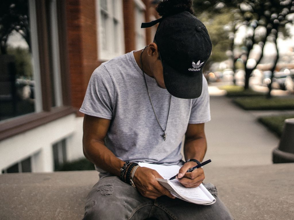 Young person wearing gray jeans, a gray t-shirt, a black ballcap, a long necklace, bracelets, and a watch. They are facing the camera with their head down, looking at a notebook they're writing in. They are sitting on a concrete wall. The background is blurry; on the left is a red brick building, and on the right are trees planted along the sidewalk.