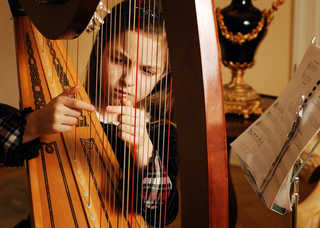 In this image taken indoors with warm lighting, a young child with long sandy blond hair sits behind a wooden harp and strums the instrument's strings. Beside the child and the harp is a metal music stand holding sheet music. Photo by <a href="https://unsplash.com/@heidiyanulisphotography?utm_source=unsplash&utm_medium=referral&utm_content=creditCopyText">Heidi Yanulis</a> on <a href="https://unsplash.com/?utm_source=unsplash&utm_medium=referral&utm_content=creditCopyText">Unsplash</a>