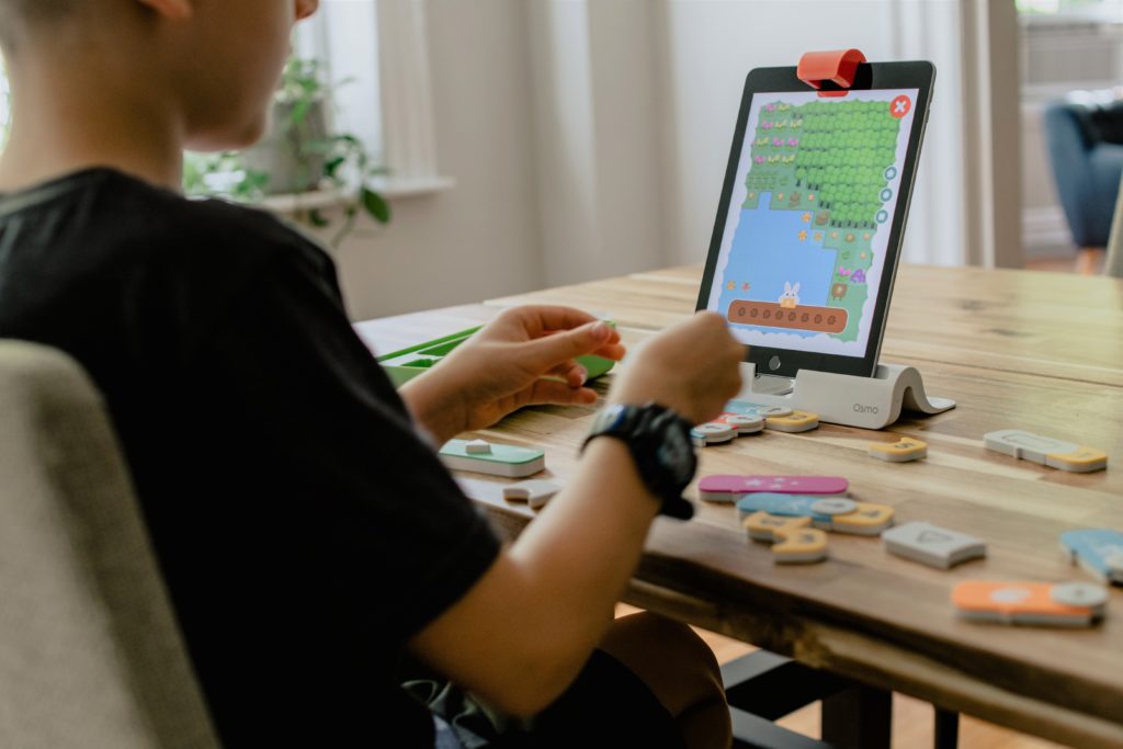 Young person wearing a black short-sleeved t-shirt with a black sports wristwatch. The young person is seated at a wood table indoors and is practicing a shapes learning activity on a computer tablet with physical shapes. In the background there is a window with natural light and a potted plant on the windowsill. Image credit: Kelly Sikkema.