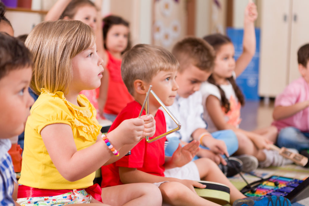 A group of pre-K students sit together in a classroom and play musical instruments, including a triangle and xylophone.