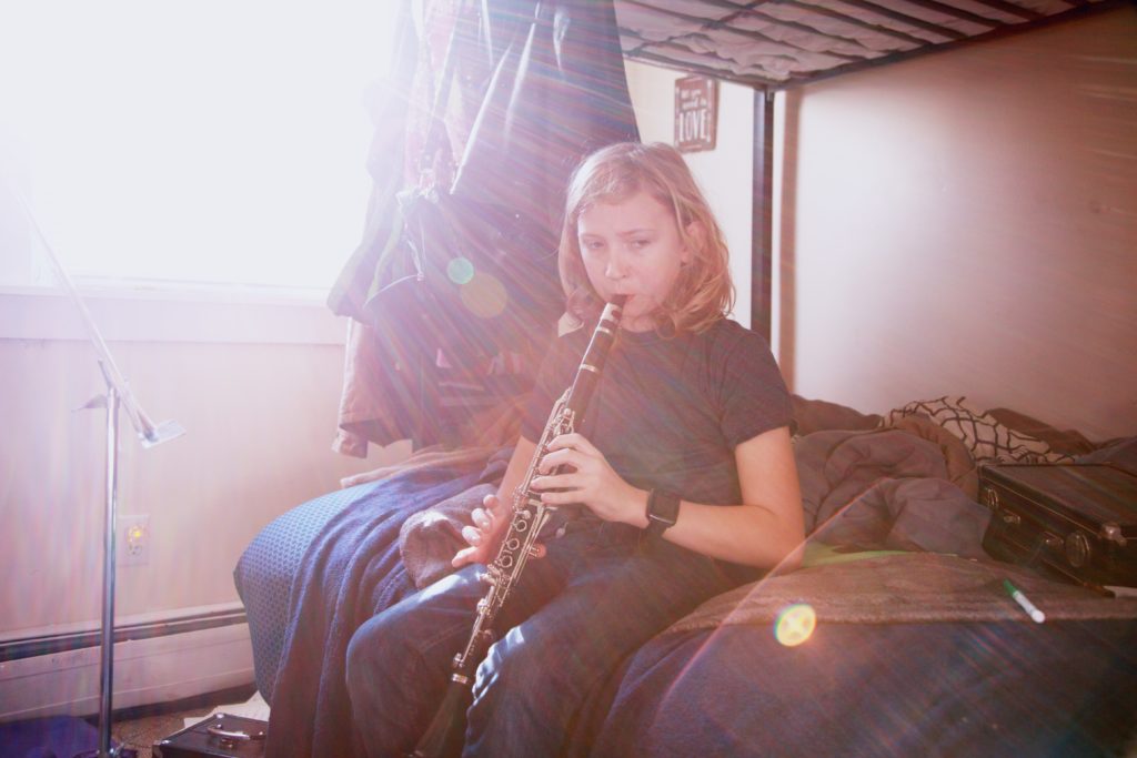 A child with shoulder-length sandy blond hair sits alone on the bottom bunk of a bunk bed and plays clarinet. The bed is unmade, and the child is wearing a short-sleeved t-shirt and denim jeans and is alone in the room. The child is in the center of the photo, with the clarinet case tossed behind them on the bed and a metal music stand to the left of them. Natural light filters into the room from the left side of the photo and creates a lens flare. Photo by <a href="https://unsplash.com/@sharonmccutcheon?utm_source=unsplash&utm_medium=referral&utm_content=creditCopyText">Sharon McCutcheon</a> on <a href="https://unsplash.com/s/photos/student-music?utm_source=unsplash&utm_medium=referral&utm_content=creditCopyText">Unsplash</a> 